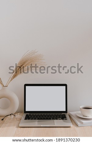 Laptop with blank copy space screen on table with pampas grass bouquet in vase, glasses, magazines and coffee cup. Minimalist home office workspace. Mockup template.