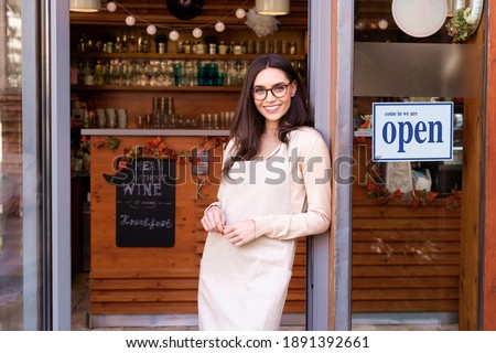 Shot of smiling young cafe show owner woman standing with arms crossed in the doorway. Open sign on the glass door.