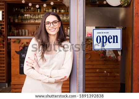 Shot of smiling young cafe show owner woman standing with arms crossed in the doorway. Open sign on the glass door.