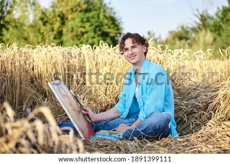 Young male artist, wearing blue shirt and torn jeans, sitting on wheat field, drawing on canvas. Painting workshop in rural countryside. Artistic education concept. Outdoors leisure activities.