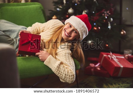 Waist up portrait of joyful girl in Santa hat relaxing near Christmas tree and holding present