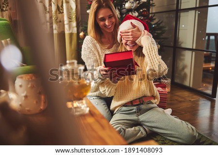 Joyful teenage girl in Santa hat is holding box while her mother is closing her eyes with hand at home