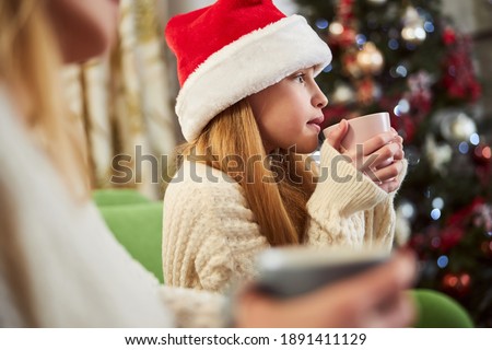 Pretty girl in Santa hat is enjoying hot drink while celebrating holidays with family indoors