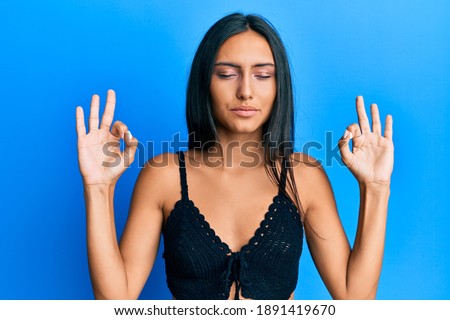 Young brunette woman wearing summer top over blue background relax and smiling with eyes closed doing meditation gesture with fingers. yoga concept. 