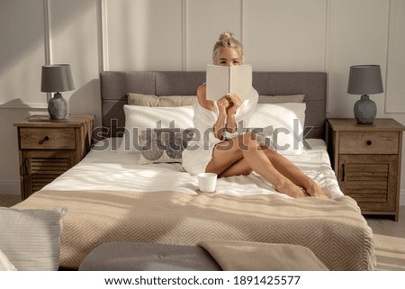 White cozy bed and a beautiful blonde girl in a white shirt with a book, a cup with a drink. The concept of a cozy holiday, self-isolation at home