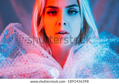 Beauty art portrait. Plastic pollution. Waste reuse. Save planet. Worried woman wrapping dress bubbles polyethylene film looking at camera blue pink neon light background.