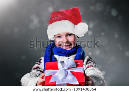 Cute child Santa holding red Christmas box with silky ribbon on gray background
