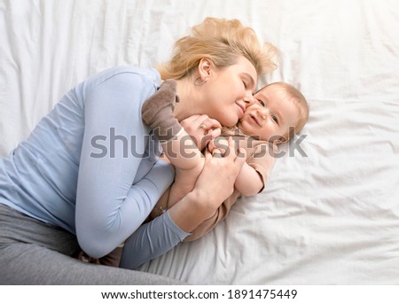 Love and tenderness, motherhood concept. Loving blonde mother caressing her cute baby boy, lying together on bed, top view. Young caucasian woman bonding with little kid at home, copy space Royalty-Free Stock Photo #1891475449