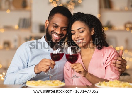 Cheers. Beautiful lovely young african american couple sitting at table in restaurant or at home, drinking red wine, holding glasses during romantic dinner. Smiling black man embracing his happy woman Royalty-Free Stock Photo #1891477852