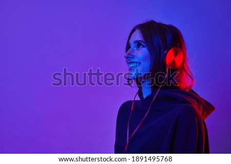 Fashion hipster girl smiles and wear headphones listening to music over color neon background at studio. Royalty-Free Stock Photo #1891495768