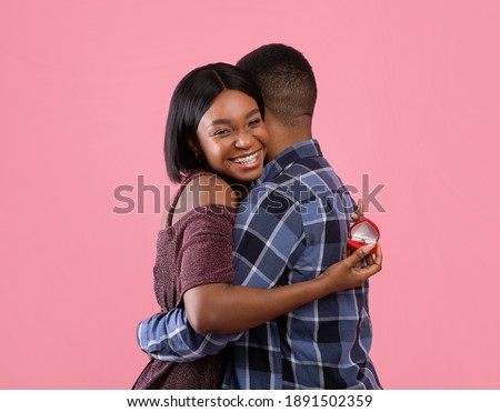Happy black woman hugging her fiance and holding engagement ring on pink studio background. Passionate lady accepting marriage proposal on Valentine's Day, feeling happy and loved