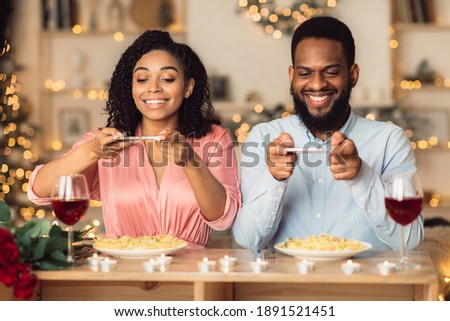 Excited young african american couple sitting in restaurant and taking pictures of food with mobile phones for social media, foodie bloggers celebrating St Valentine's Day together