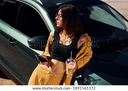 With smartphone. Young fashionable woman in burgundy colored coat at daytime with her car.