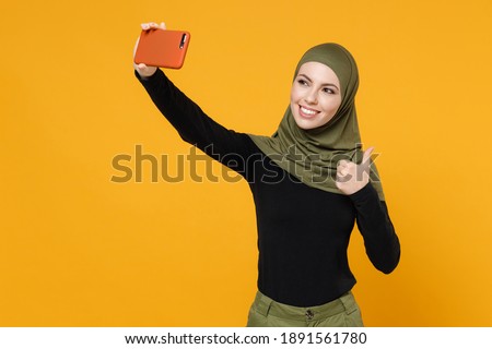 Smiling young arabian muslim woman in hijab black green clothes doing selfie shot on mobile phone showing thumb up isolated on yellow background studio portrait. People religious lifestyle concept