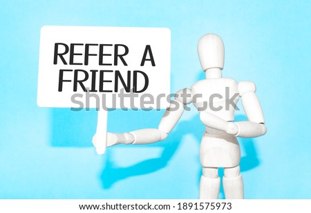 The wooden man holds a white sign with the text REFER A FRIEND in his hands. The content of the lettering has implications for business concept and marketing.