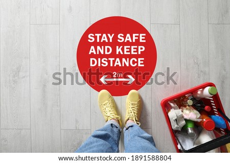 Woman near sign STAY SAFE AND KEEP DISTANCE in supermarket, top view. Coronavirus pandemic
