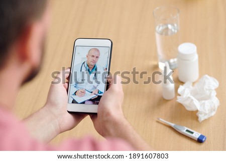 Mature male doctor on smartphone screen making notes and giving medical recommendations to young sick man describing his symptoms