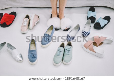 Woman trying on cosy fluffy open toed house slippers among different pairs of moccasins sneakers slippers beside on grey floor