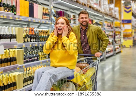 couple shopping together in grocery supermarket, man carry his redhead girlfriend on cart, they have fun, enjoy time, woman is surprised happy