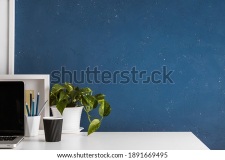 White table against blue wall with home office supplies, plant, laptop and coffee cup. 