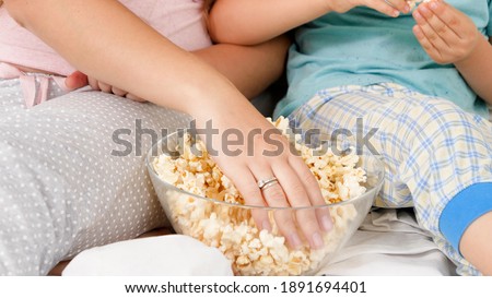 Closeup of mother and son in pajamas taking popcorn from big glass bowl while watching cartoons or movie in bed. Concept of cheerful children and family happiness