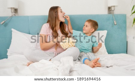 Little toddler boy with young mother in pajamas lying in bed on weekend and eating popcorn from big bowl. Concept of cheerful children and family happiness