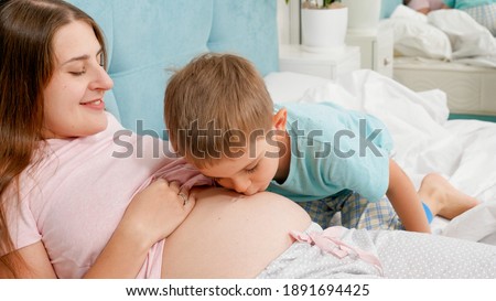 Cute adorable boy kissing and listening to big belly of his pregnant mother lying on bed. Concept of loving children and family happiness expecting baby.
