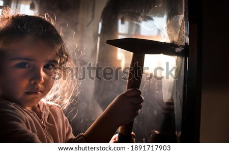 Cute little girl standing in front of a TV with broken screen holding a hammer. Home insurance concept. Child smashed led TV screen with a hammer