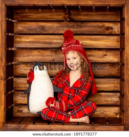 A girl in Christmas pajamas in a hat and mittens hugs a stuffed penguin toy on a wooden background. Happy New Year
