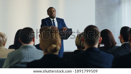 Male African American business speaker standing at the lecturn at the conference and interacting with people in the hall during speech while someone from the audience rising hand and asking the Royalty-Free Stock Photo #1891799509
