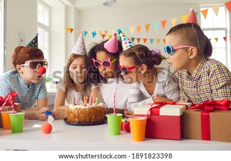 Happy Birthday. Group of diverse little children in paper hats and party sunglasses all together blowing candles on cake standing at festive table during fun celebration at home