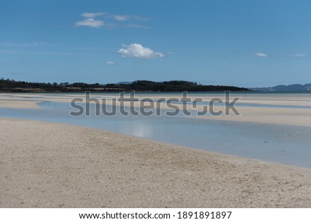 Calm water on white brown sand with distant mountains in the background and a blue clear sky above