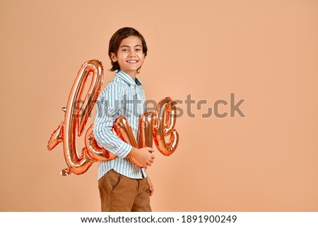 A nice Caucasian boy in a striped shirt holding a balloon with the words "love", isolated on a delicate peach background. Valentine's Day.