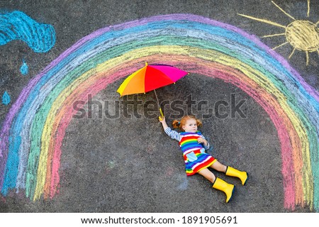 Happy little toddler girl in rubber boots with rainbow sun and clouds with rain painted with colorful chalks on ground or asphalt in summer. Cute child with umbrella having fun. creative leisure .