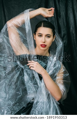 Beauty care. Plastic surgery. Anti aging procedure. Confident woman with perfect face skin night makeup red lips posing in wrinkled transparent polyethylene film veil isolated on dark background.