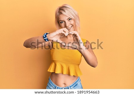 Young blonde girl wearing casual clothes smiling in love doing heart symbol shape with hands. romantic concept. 