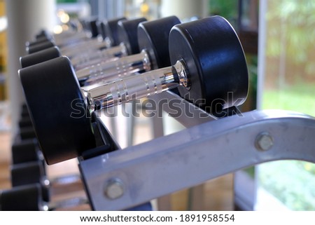 A close-up picture of steel dumbbells with black rubber protection lining up in rows on a metal rack in a gym by a glass window. 