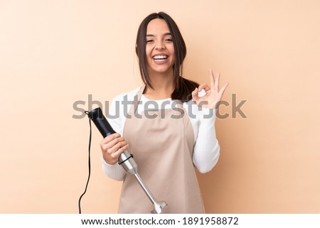 Young brunette girl using hand blender over isolated background showing an ok sign with fingers