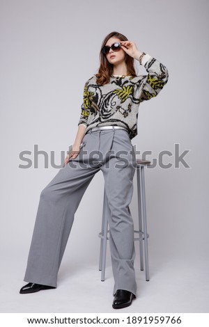 Fashion photo of a beautiful elegant young woman in a pretty  pants, trousers, blouse, accessories, boots, stylish sunglasses posing over white, soft gray background. Brown hair. Studio Shot, portrait