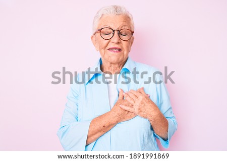Senior beautiful woman with blue eyes and grey hair wearing glasses smiling with hands on chest with closed eyes and grateful gesture on face. health concept. 