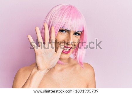 Young man wearing woman make up wearing pink wig showing and pointing up with fingers number five while smiling confident and happy. 