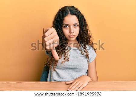 Teenager hispanic girl wearing casual clothes sitting on the table looking unhappy and angry showing rejection and negative with thumbs down gesture. bad expression. 