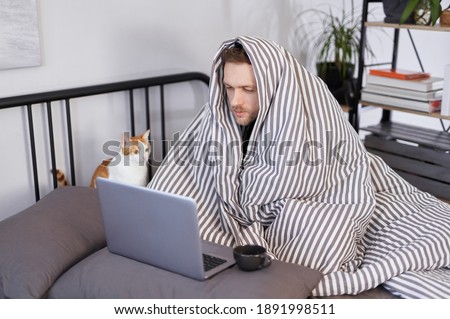 Bearded young man using laptop in bed full covered with blanket and white ginger cat near him. Concept of distant work from home office or online studying while feeling sick Royalty-Free Stock Photo #1891998511