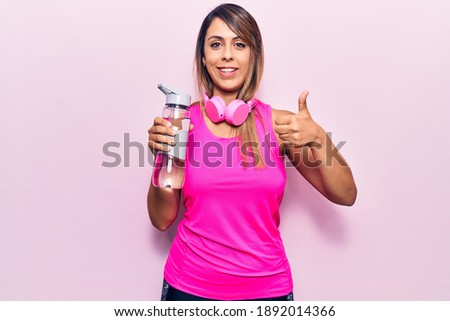 Young beautiful woman wearing sportswear drinking bottle of water using headphones smiling happy and positive, thumb up doing excellent and approval sign 