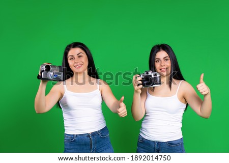 Twin girls posing with vintage camera and vintage video camera and showing class on green background.