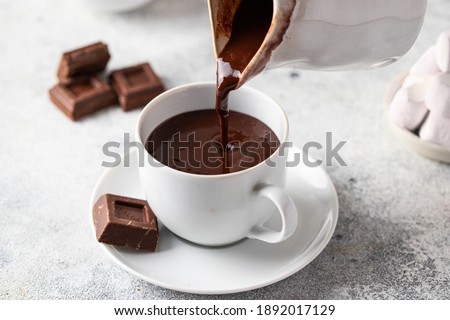 Pouring hot chocolate  from a jug into the cup on white grey background. Winter drink concept.