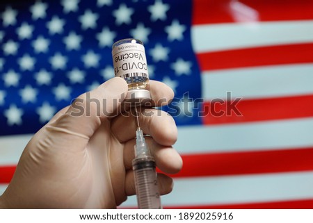 Close up hand of a medical staff holding a Covid-19 vaccine and syringe against American flag background. Coronavirus prevention, immunization and treatment.