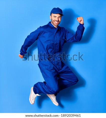 Young handsome hispanic man wearing painter uniform and cap smiling happy. Jumping with smile on face doig winner sign with fists up over isolated blue background