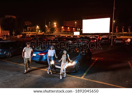 Full length shot of three diverse young friends looking at camera while standing by the car parked in front of a big screen, ready to watch a movie at drive in cinema. Friendship, leisure concept