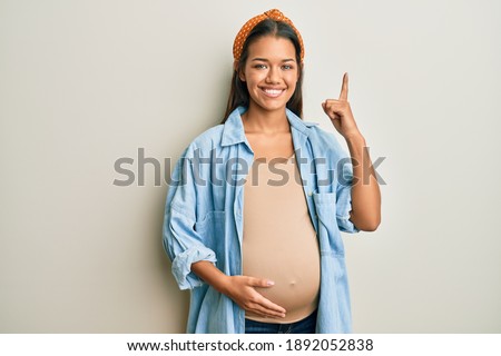 Beautiful hispanic woman expecting a baby, touching pregnant belly with a big smile on face, pointing with hand finger to the side looking at the camera. 